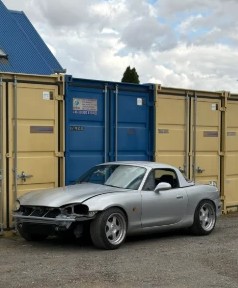 The MX-5 parked by shipping containers, front bumper dented